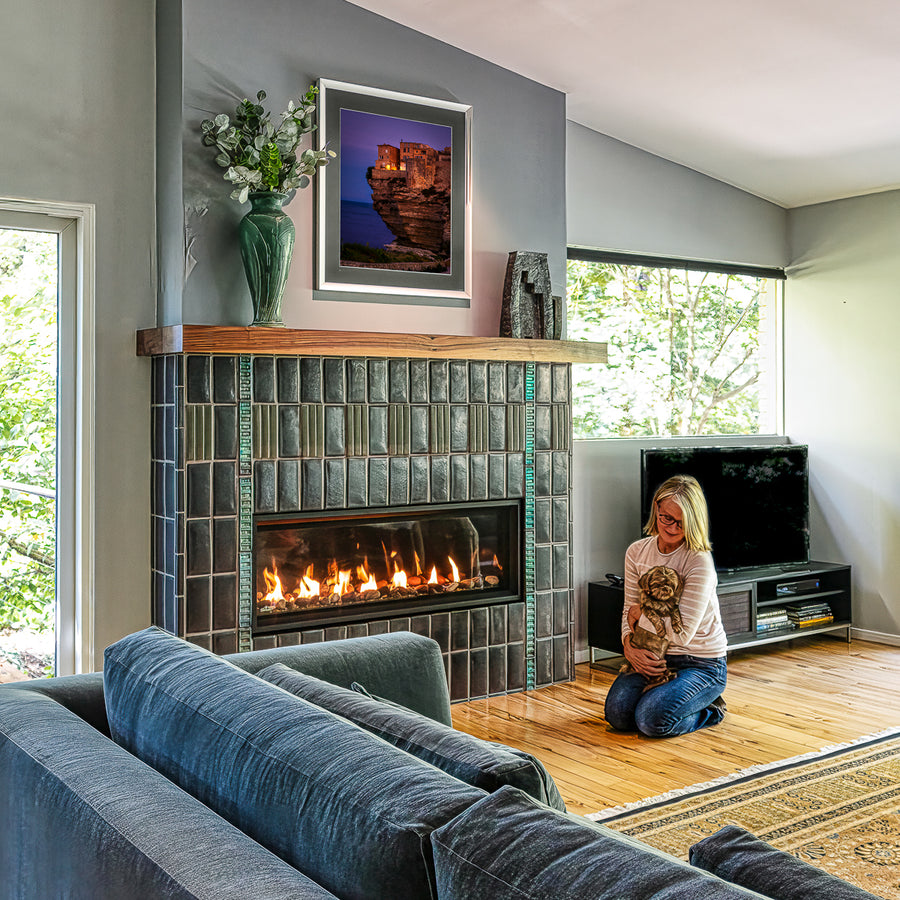 Angled view of a custom Pewabic fireplace set inside of a Midcentury-style home. There is a person holding a small dog near the lit fireplace. The windows on either side of the fireplace bring out the vibrant shine of the tilework.
