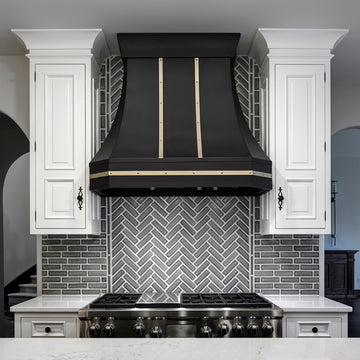 Light Gray Modern Herringbone pattern tiles line the wall behind a stainless steel stove with a large matte black hood. Ornate white cabinets flank the tile on either side.