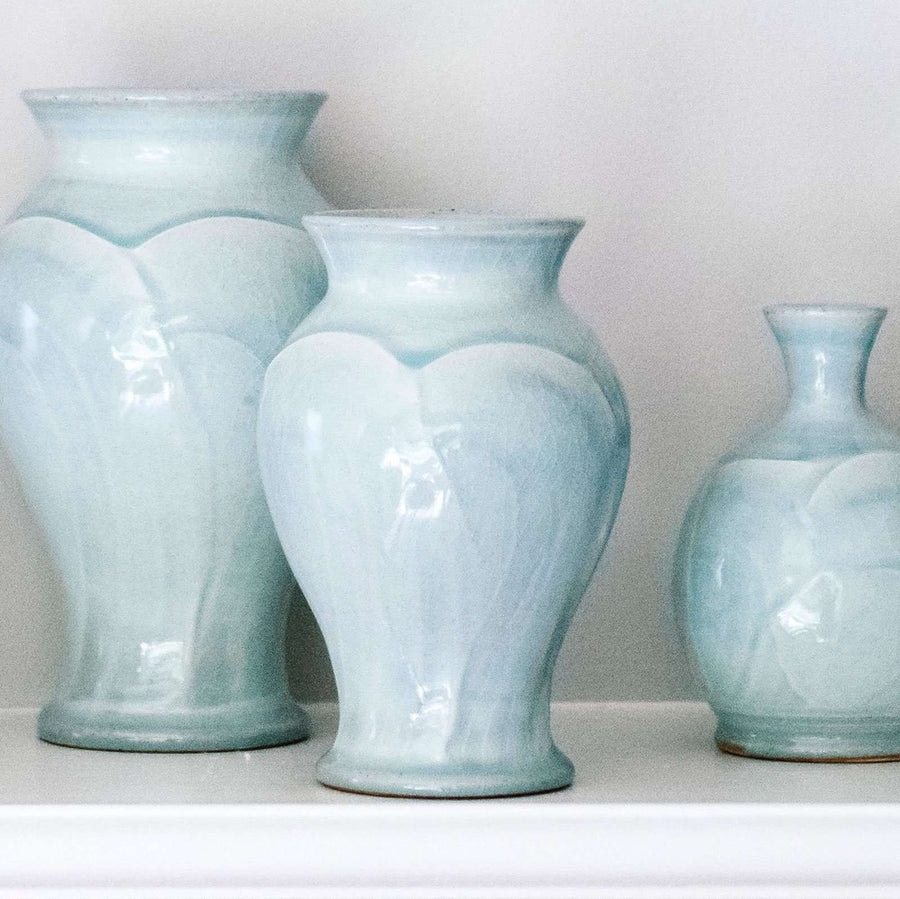 Three vases in various shapes sit on a white shelf- they all feature the glossy pale blue-gray Frost glaze. A close-up of the Frost glaze shows crazing on its surface- this gives the appearance of tiny cracks on the glaze's surface.