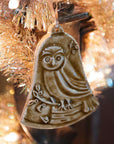 This bell-shaped ornament features a wide-eyed owl sitting on a fallen tree trunk, branches covered in leaves and berries surround it.