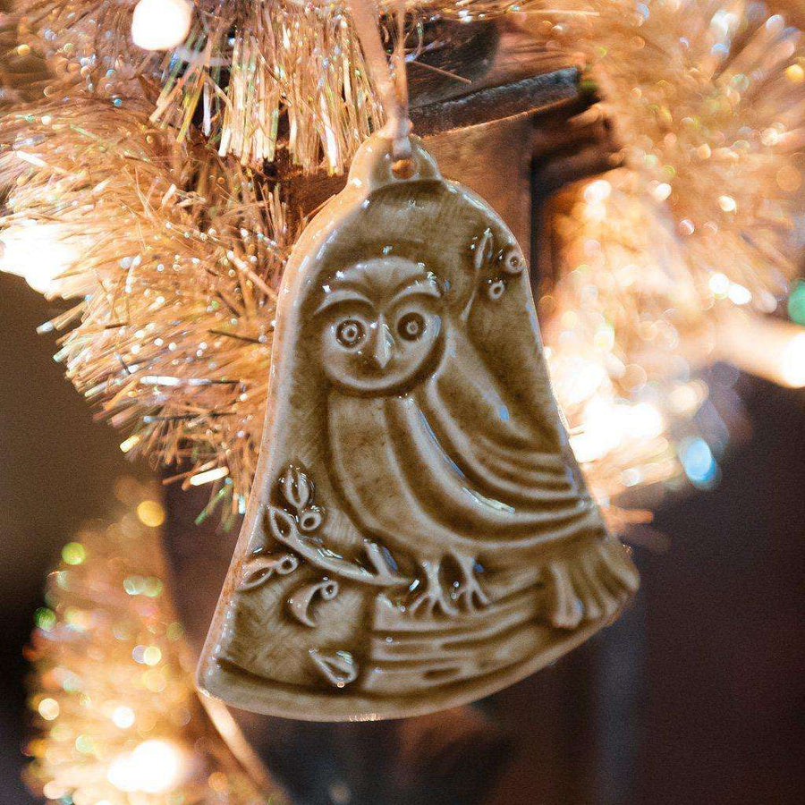 This bell-shaped ornament features a wide-eyed owl sitting on a fallen tree trunk, branches covered in leaves and berries surround it.
