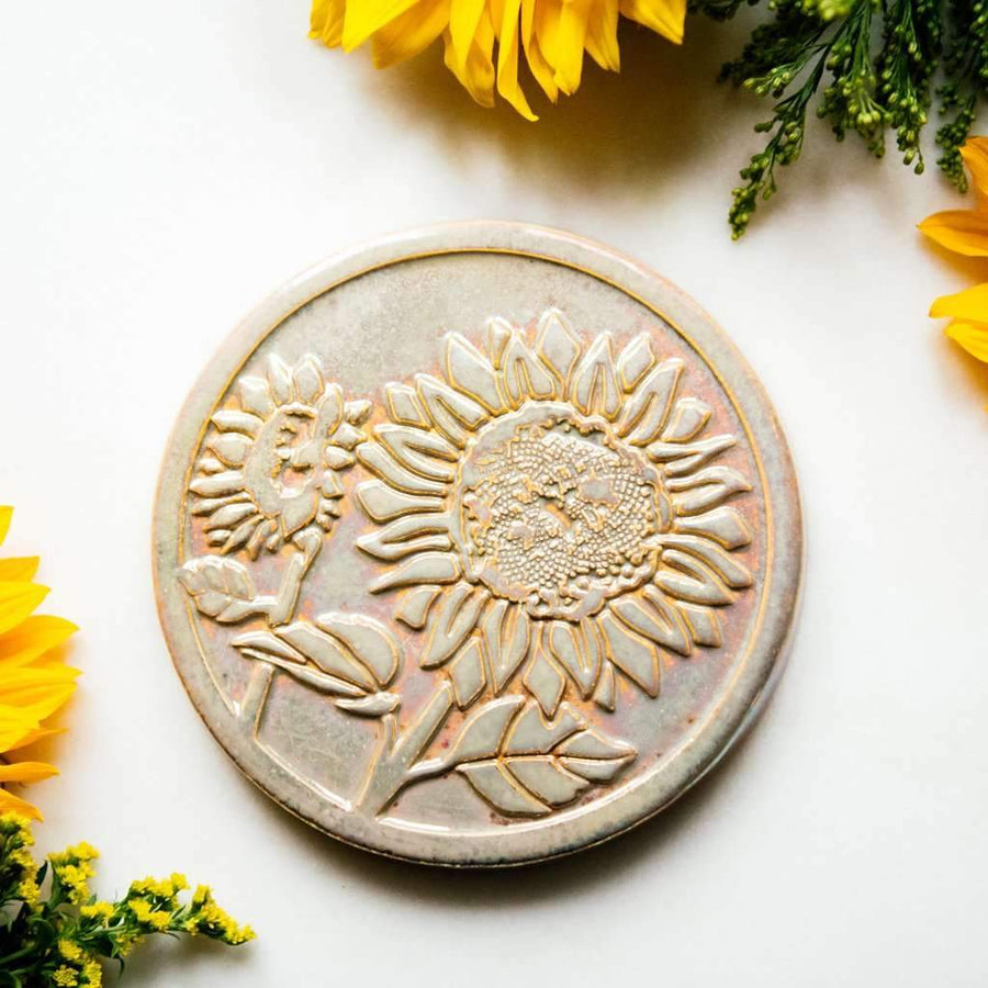 The Sunflower Trivet Tile features two sunflowers growing up into the circular tile with leaves and a bit of stalk in view. One sunflower is facing forward while the other is slightly smaller and facing to the left.