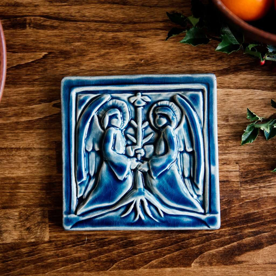 The square tile shows two angels in flowing robes with large feathered wings face each other, each has their hands grasping a bare tree growing between them. This tile features the glossy deep blue Ocean glaze.