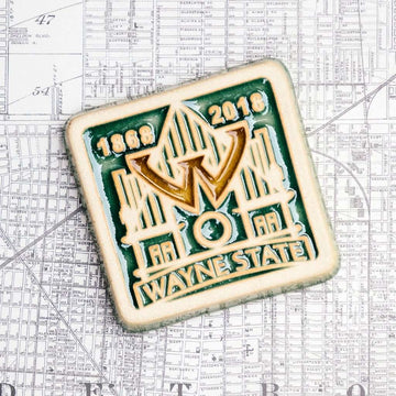 The Wayne State University tile is displayed on a map of Detroit. The tile boasts a prominent W in the center of the design that is hovering over a simple line drawing of the Old Main. On the bottom of the tile "Wayne State" is written in bold lettering and the dates 1868 and 2018 are displayed across the top- in celebration of Wayne State's 150 years as a learning institution. This whole design is depressed into a flat tile. The depressions hold glossy green and gold glaze.