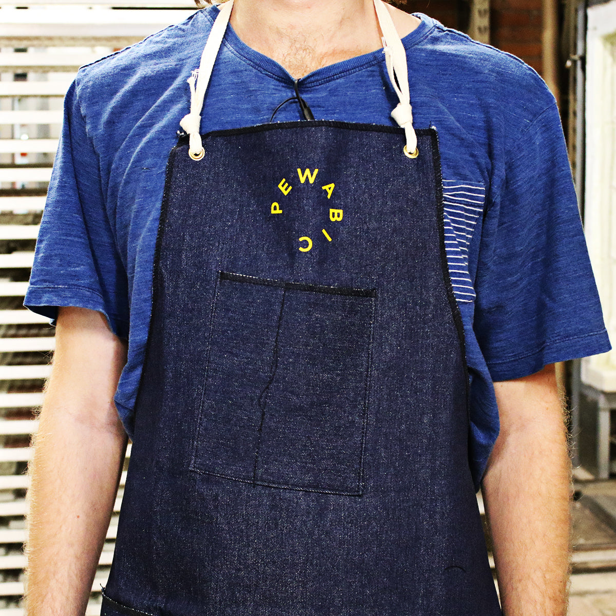 The Pewabic Apron is a dark denim wash with the Pewabic logo in bright yellow in the center of the chest.