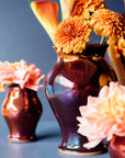 A copper glazed vase holds many petalled flowers. The surface on the glaze is so smooth, it is reflective like a mirror.