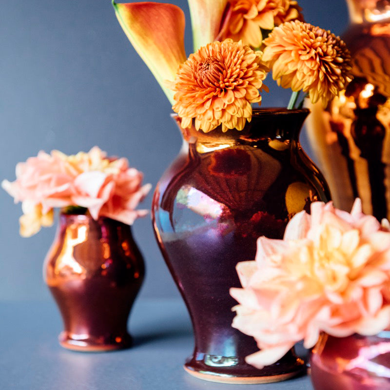 A copper glazed vase holds many petalled flowers. The surface on the glaze is so smooth, it is reflective like a mirror.