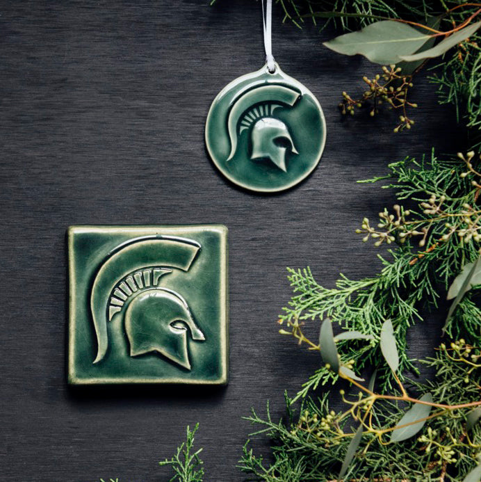 The MSU Tile and Ornament are sitting next to each other. Both pieces feature the same Spartan helmet design.