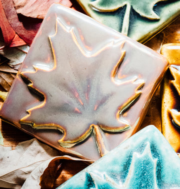 The iridescent maple leaf tile lays on a bed of fall foliage. The surface of the embossed maple leaf design is smooth and metallic.