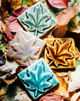 All four colors of the maple leaf embossed tile are resting on a bed of fall leaves. Each maple leaf design covers the entire surface of the tile. 