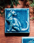 A tile sits on a wooden table surrounded by greenery. The matte blue embossed tile shows a sleek cat sitting up straight with its head turned backward towards its flicking tail.