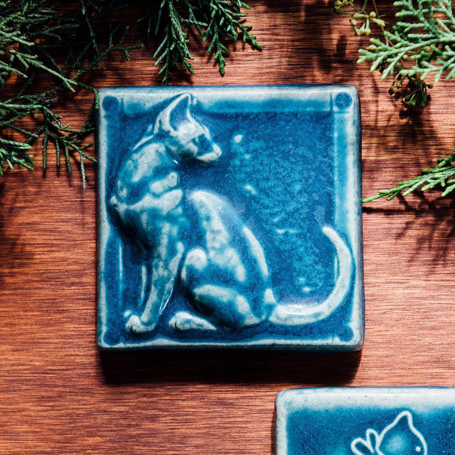 A tile sits on a wooden table surrounded by greenery. The matte blue embossed tile shows a sleek cat sitting up straight with its head turned backward towards its flicking tail.