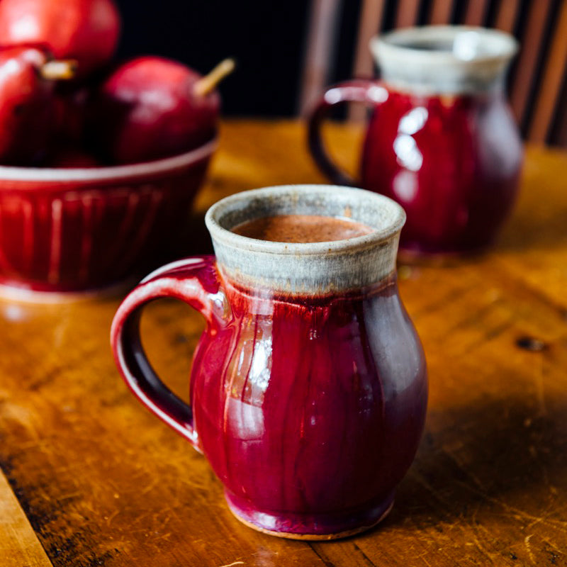 Two mugs sit on a warn wooden table. A Winterberry bowl sits behind them holding red pears. 