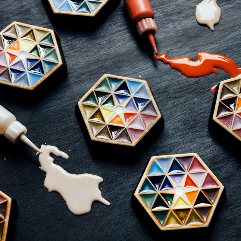Many Hex Paperweights sit on a table. Small bulbs with long skinny applicators drip their colorful glazes onto the table around the tiles.