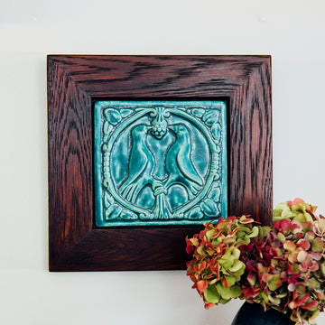 The Lovebirds Tile has a high relief design showing two birds facing one another on a tree branch while pecking at the same berry. There is an ornate border design around it including leaves and twirled lines. The tile features the matte turquoise Pewabic Blue glaze which beautifully offsets the deep reddish brown hues of the oak wood frame.