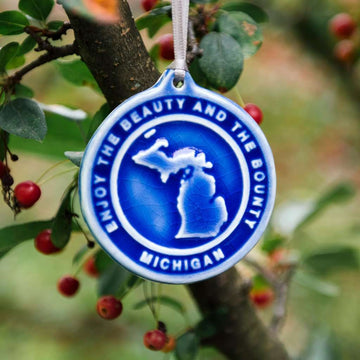 The round Michigan Ornament features the state's two peninsulas surrounded by a border with the words "enjoy the beauty and the bounty, Michigan" written around the circular edge.