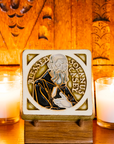 The Solanus Casey Tile features a line drawing of Solanus Casey from mid-chest up wearing glasses and a dark brown cloak while holding a Bible. The words "Blessed Solanus Casey" are curved around him, creating a halo-like circle. This hand painted tile features a Honey Gloss Glazed background with dark brown on his clothing and gray on his beard.