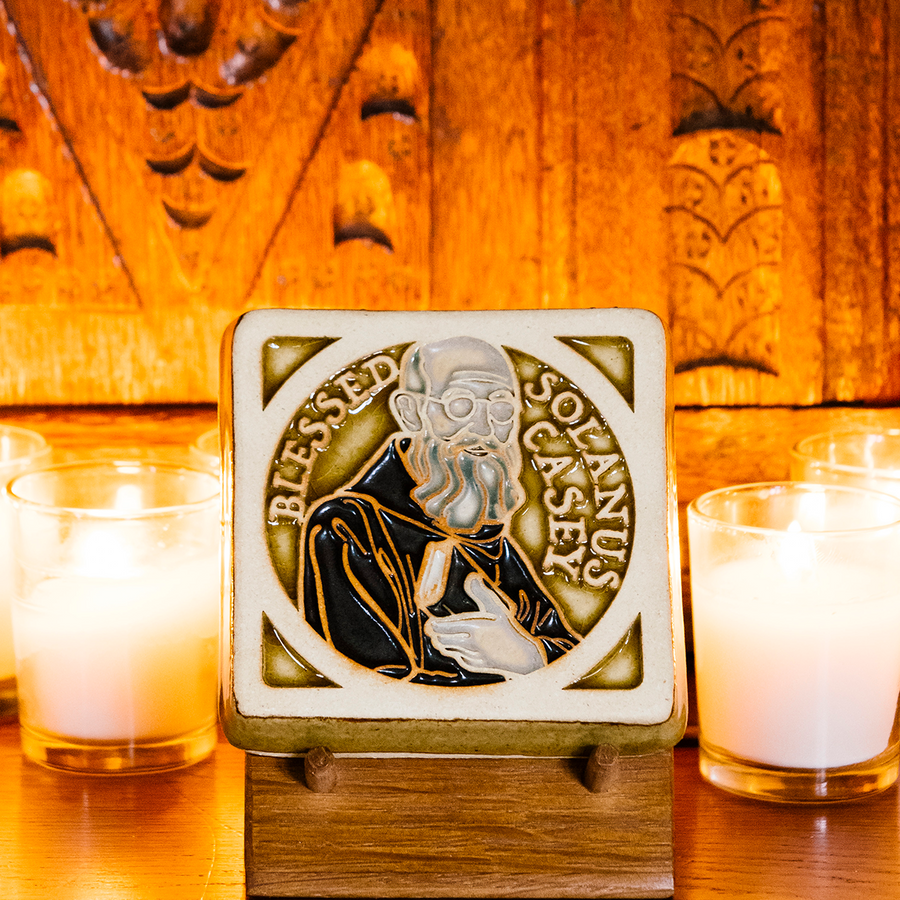 The Solanus Casey Tile features a line drawing of Solanus Casey from mid-chest up wearing glasses and a dark brown cloak while holding a Bible. The words "Blessed Solanus Casey" are curved around him, creating a halo-like circle. This hand painted tile features a Honey Gloss Glazed background with dark brown on his clothing and gray on his beard.