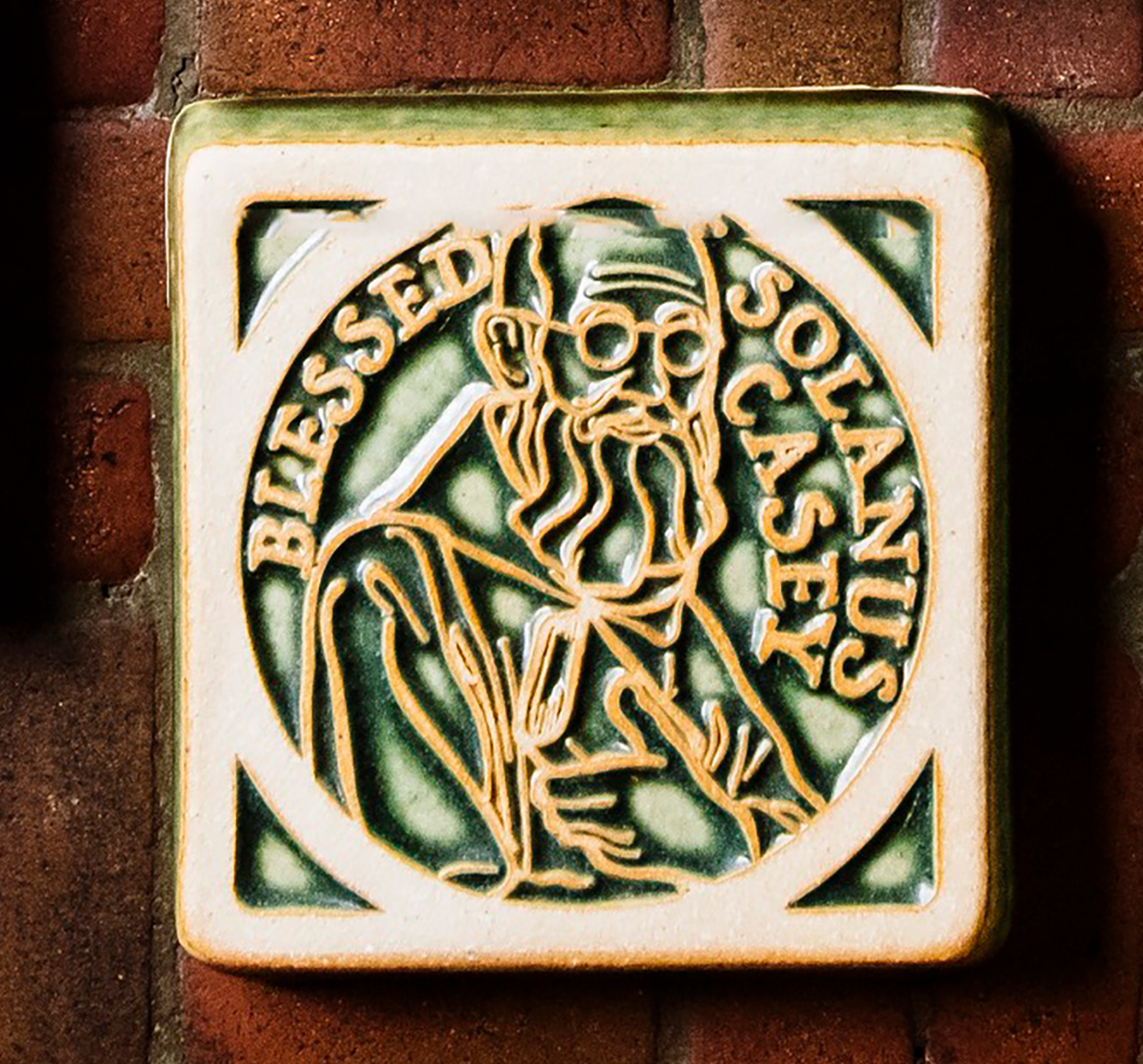 The Solanus Casey Tile features a line drawing of Solanus Casey from mid-chest up wearing glasses and a cloak while holding a Bible to his chest. The words "Blessed Solanus Casey" are curved around him, creating a halo-like circle. This tile is glazed in a glossy green color with the raised design being scraped giving it a creamy white hue.