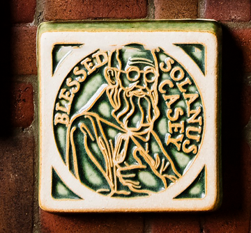 The Solanus Casey Tile features a line drawing of Solanus Casey from mid-chest up wearing glasses and a cloak while holding a Bible to his chest. The words "Blessed Solanus Casey" are curved around him, creating a halo-like circle. This tile is glazed in a glossy green color with the raised design being scraped giving it a creamy white hue.
