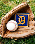 A leather baseball mitt is nestled in bright, green grass. Inside the mitt lays a baseball and our 4x4" Tigers Old English "D" Tile in a Royal Blue and burnt orange "Natural" border. The Old English "D" logo is also outfitted in burnt orange, allowing it to stand out against its bright backdrop.
