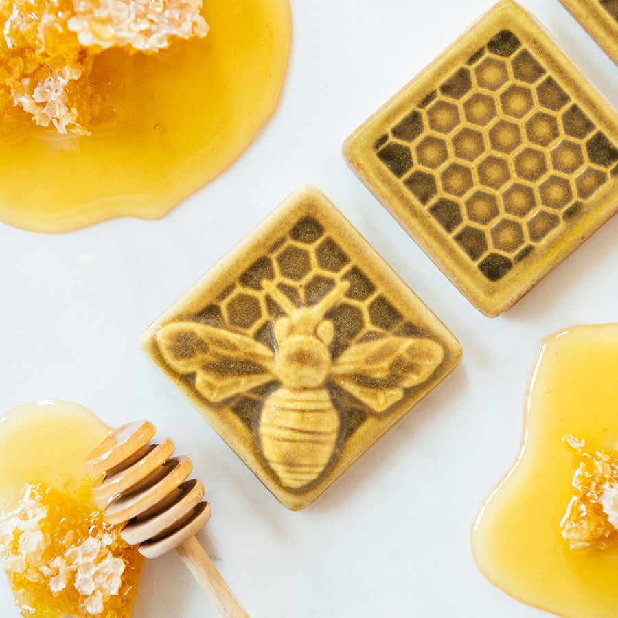 The Honeybee Tile features a large honeybee with stripes and detailed segmented wings. The bee sits on a honeycomb patterned background. These tiles features the matte golden yellow Mustard glaze.