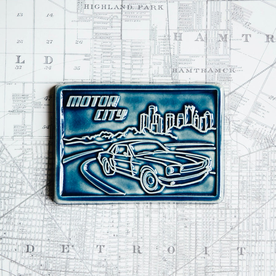 The Motor City Postcard Tile features a line drawing of a classic car racing down the road toward you with the Detroit skyline in the background. In the sky are the words "Motor City" in bold writing. This tile is in the glossy deep blue Ocean glaze.