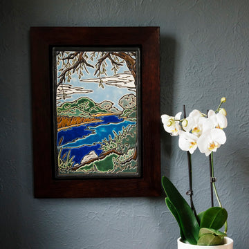 The Lake Okonoka Tile features a snapshot of the lake with bushes lining its banks and a large willow tree in the foreground that frames it. This gives the impression that you are seated on the grass under the tree next to the serene water. This tile is hand painted in blues and greens which are offset by the beautiful deep reddish brown oak wood frame.