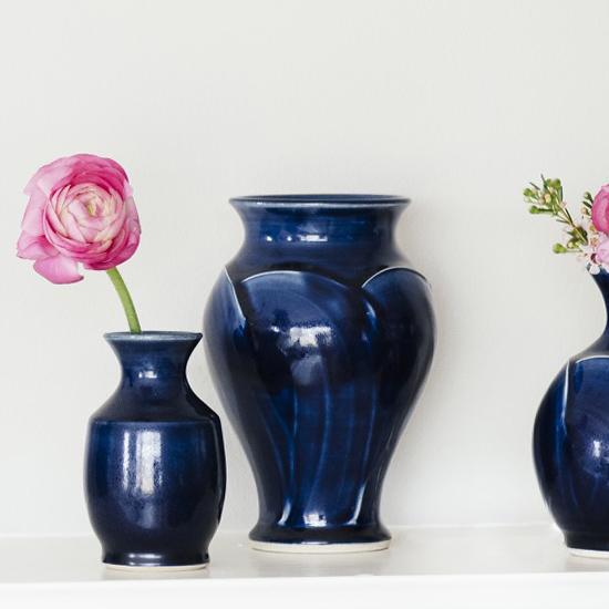 Multiple vases stand together holding delicate flowers. The vases feature the glossy deep dark blue Midnight glaze.