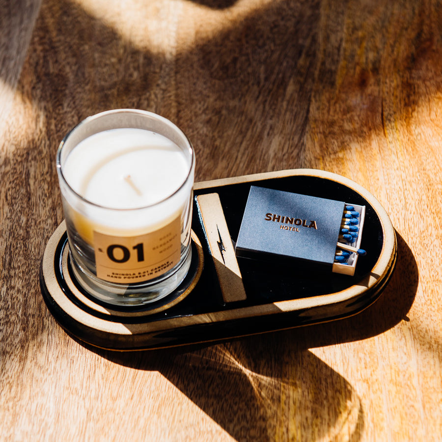 The Shinola candleholder is a large oval tile in a dark glossy glaze. There is an inset border around the edge of the tile, making it closer to a tray. Down the center of the tile is an short thick line design with the Shinola lightening logo inside of it - this line divides the tile into to sections. One section only has the smooth glaze while the other section has a raised circle where you can place your round-bottomed Shinola candle.