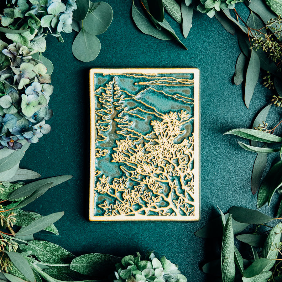 This Mountain View Tile features a a large flowery bush in the foreground with a tall pine tree behind it. In the distance, craggy mountains sit under a cloudy sky.  