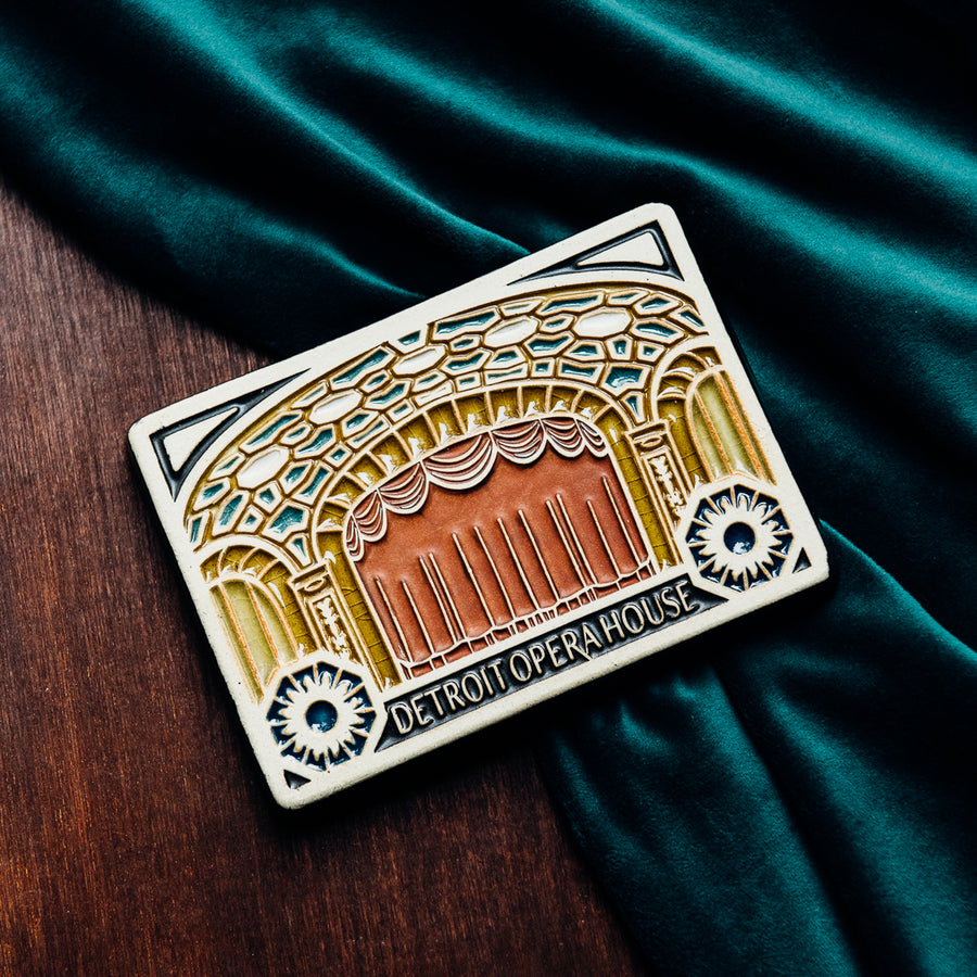 The Michigan Opera Theatre Postcard Tile design features the stage of the theatre with the curtains closed. The ornate pillars and ceiling are punctuated with blues and whites. Under the stage reads the words "Detroit Opera House" with two geometric floral designs on either side.