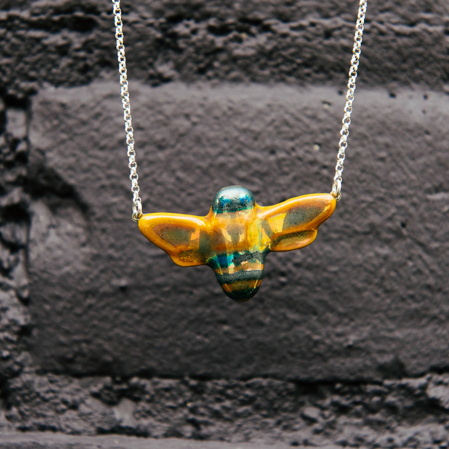 The bee necklace is held up against a black painted brick wall.