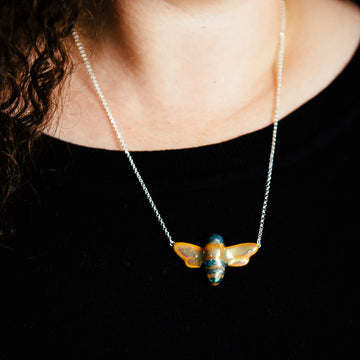 The Bee Necklace features a high-relief bee with wings outstretched. The hand-painted iridescent glaze gives it black stripes and a black head while the rest of the bee features the gold Blush Iridescent glaze. The bee is smooth and shiny and its wings are attached to the silver chain.