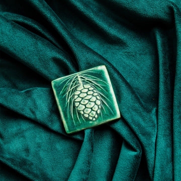 The ceramic Pinecone Tile has a high relief design with one large pinecone in the center and long spindly pine needles framing it. This tile features the matte blueish-green Pewabic Green glaze.