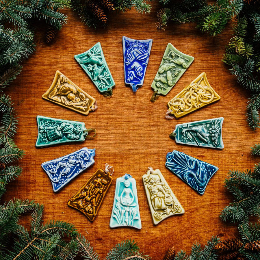 A colorful ceramic 12 Days of Christmas Ornament Set is splayed in a perfect circle with boughs of greenery surrounding them.