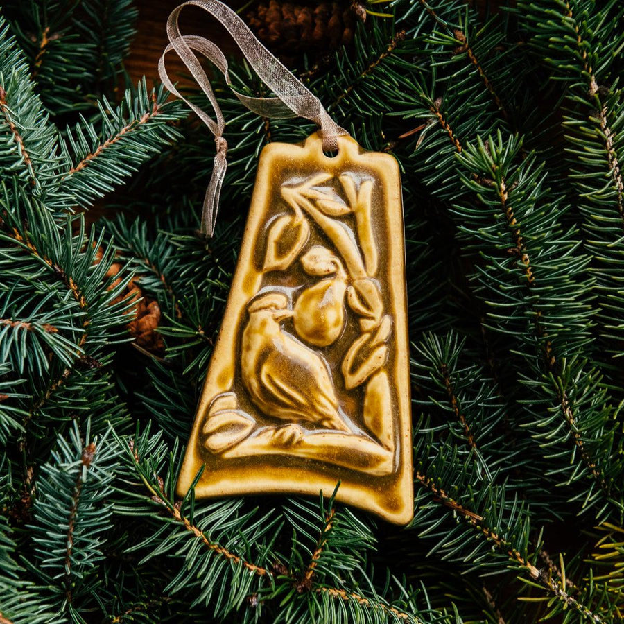 The bell-shaped Partridge in a Pear Tree Ornament features a partridge sitting on the branches of a tree with one large pear dangling from its branches. The ornament is glazed in a creamy brown color.