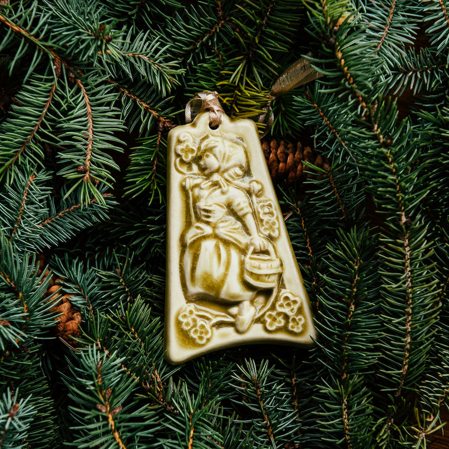 The bell-shaped Eight Maids a-Milking Ornament features a young girl wearing a Bavarian style dress and head scarf. She is carrying a wooden bucket and there are eight small flowers surrounding her. The ornament is glazed in a matte cream glaze.