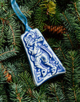 The bell-shaped Eleven Pipers Piping ornament features a man wearing a medieval costume with a feather in his hat. He is playing a long conical pipe and there are eleven musical notes surrounding him. The ornament is glazed in a matte blue glaze.
