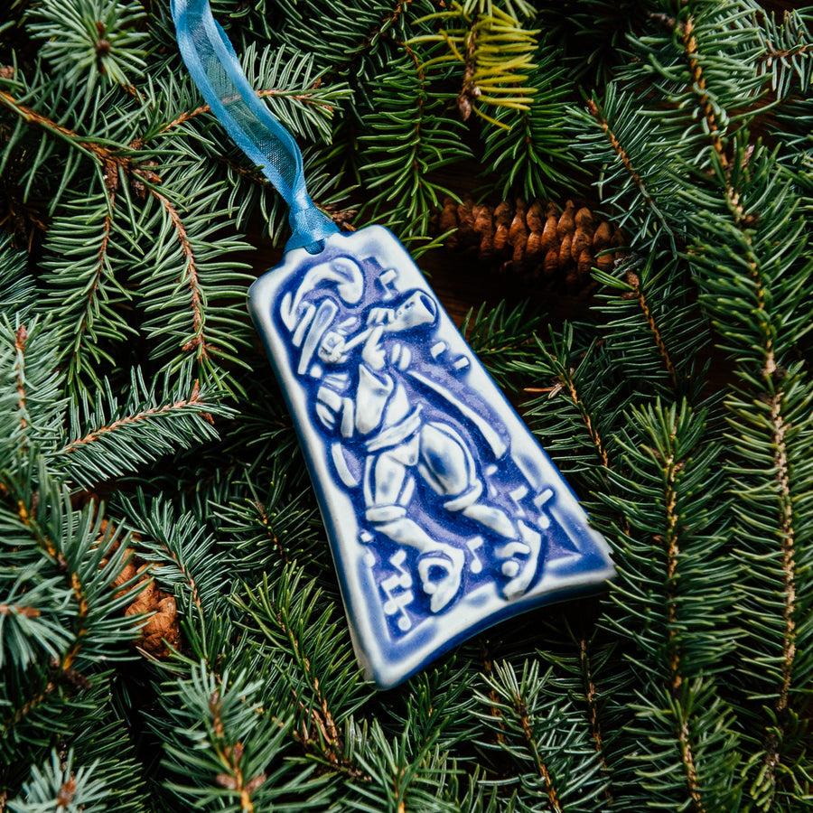 The bell-shaped Eleven Pipers Piping ornament features a man wearing a medieval costume with a feather in his hat. He is playing a long conical pipe and there are eleven musical notes surrounding him. The ornament is glazed in a matte blue glaze.