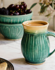Classic Mug in Pewabic Green in the foreground filled to the brim with coffee and cream. In the background, there are two Small Pewabic Classic Bowls stacked on top of one another with blackberries and dark, red cherries. There is a Birch Petite Mug in the corner of the photo holding seeded  eucalyptus. A pewter tray of shortbread cookies are coming into frame on the bottom-left.