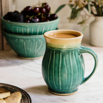 Classic Mug in Pewabic Green in the foreground filled to the brim with coffee and cream. In the background, there are two Small Pewabic Classic Bowls stacked on top of one another with blackberries and dark, red cherries. There is a Birch Petite Mug in the corner of the photo holding seeded  eucalyptus. A pewter tray of shortbread cookies are coming into frame on the bottom-left.