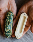 This ceramic Mezuzah features an acorn and oak leaf with the word "Peace" written in Hebrew below them. This piece is thick with an opening on the back to hold your written prayer. There are two holes at the top and bottom to make it easy to attach to your doorframe. This photo features the back of our Mezuzah in a “Leaf” glaze. 