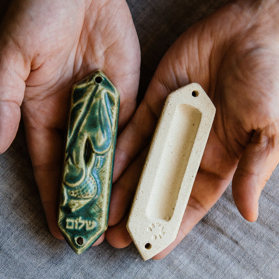 This ceramic Mezuzah features an acorn and oak leaf with the word "Peace" written in Hebrew below them. This piece is thick with an opening on the back to hold your written prayer. There are two holes at the top and bottom to make it easy to attach to your doorframe. This photo features the back of our Mezuzah in a “Leaf” glaze. 