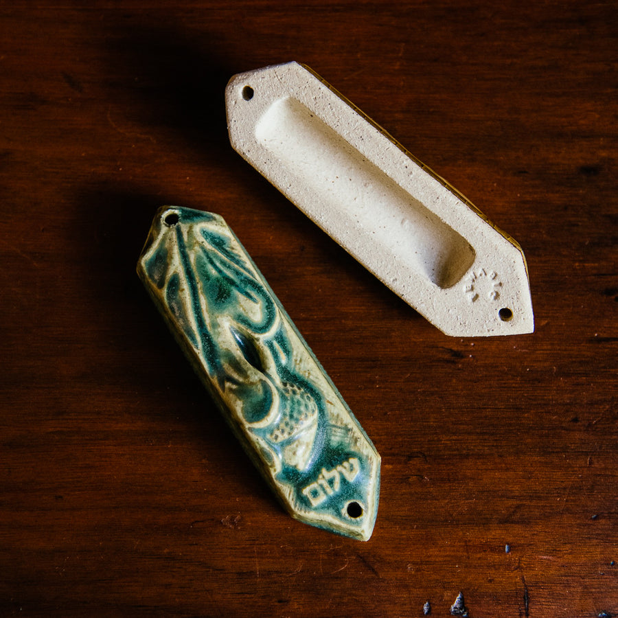 This ceramic Mezuzah features an acorn and oak leaf with the word "Peace" written in Hebrew below them. This piece is thick with an opening on the back to hold your written prayer. There are two holes at the top and bottom to make it easy to attach to your doorframe. This photo features the back of our Mezuzah in a “Leaf” glaze. 