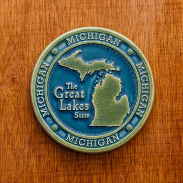 This round Michigan Trivet Tile features the outline of the state's two peninsulas including some of the larger northern islands of Michigan. To the left of the design reads "The Great Lakes State" and surrounding it, in a border, reads the word Michigan four times. This two tone Michigan tile features a matte blue background while the peninsulas and words are in a matte organic green color.