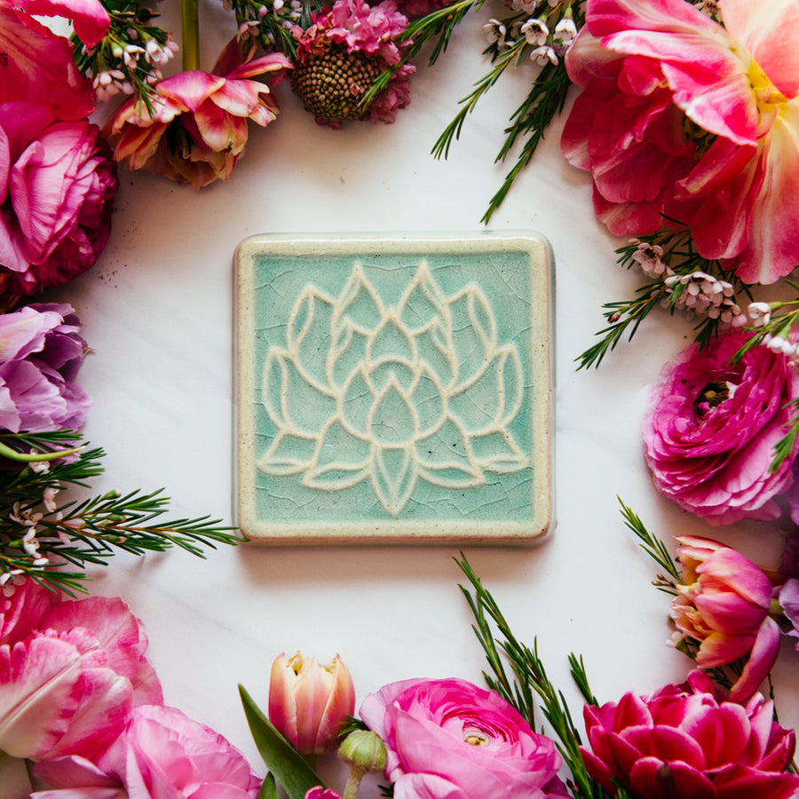 The Lotus tile features a line drawing of a blooming lotus flower, its pointed petals outstretching. It has a simple border around the design. This Lotus Tile features the glossy pale blue Celadon glaze.