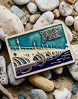 The ceramic Belle Isle Bridge Postcard tile features the Macarthur Bridge which connects Belle Isle to mainland Detroit. The words "Belle Isle" float above the bridge in the sky. This tile is hand-painted in a variety of blue glazes.