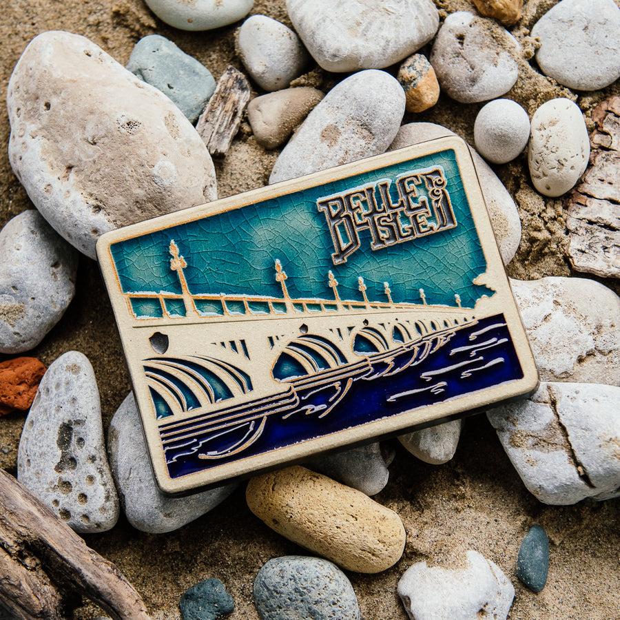 The ceramic Belle Isle Bridge Postcard tile features the Macarthur Bridge which connects Belle Isle to mainland Detroit. The words "Belle Isle" float above the bridge in the sky. This tile is hand-painted in a variety of blue glazes.