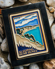 The Sleeping Bear Dunes Tile features the serene Lake Michigan and the sandy beaches and dunes covered on top by bushes and greenery. The words "Sleeping Bear Dunes" line the bottom of the design. The hand painted tile boasts blues and greens which are showcased in a black painted frame.